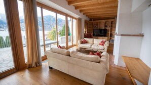 Ein geräumiges Wohnzimmer mit einer großen Couch, einem Couchtisch und einem Fernseher. Der Raum verfügt über Holzböden und eine hohe Decke mit freiliegenden Balken. Ein großes Fenster mit Vorhängen bietet Blick auf Berge und einen See.