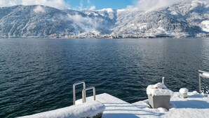 Blick auf einen See mit einem Steg, schneebedeckten Bergen und blauem Himmel.