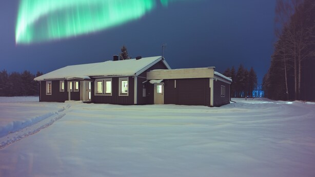 A two-story house with a snow-covered roof and a visible chimney, surrounded by a snowy landscape.