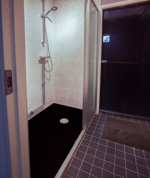 A bathroom with a shower, tiled walls, a black floor, and a visible drain. - SBC Guesthouse (Pajala)