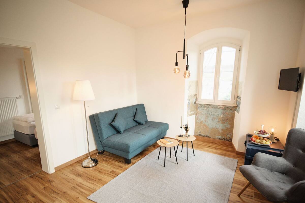 Ein modernes Wohnzimmer mit einem blauen Sofa, einem grauen Teppich, Holzböden, einem kleinen runden Tisch mit Lampe und einem an der Wand montierten Fernseher.