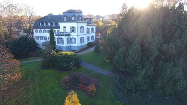 Un edificio grande y histórico con una fachada simétrica, rodeado de un jardín bien cuidado y un bosque denso.