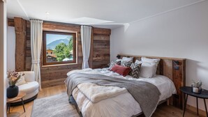 Ein Schlafzimmer mit einem hölzernen Kopfteil, einem großen Fenster mit Vorhängen, einem Bett mit vielen Kissen, einem kleinen runden Tisch und einer Vase mit Blumen.