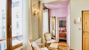 A hotel room with a wooden floor, two upholstered chairs, a small table, a bed with red bedding, a mirror, and a wooden door. - CamParis - B&B - Chambre d'hôtes (Paris)