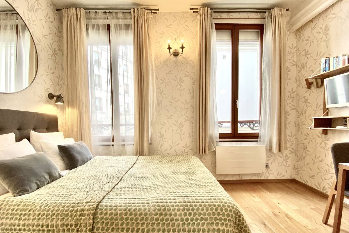 Une chambre d’hôtel avec un grand lit, une tête de lit en bois, une table de chevet, une chaise, une télévision et une fenêtre avec des rideaux.