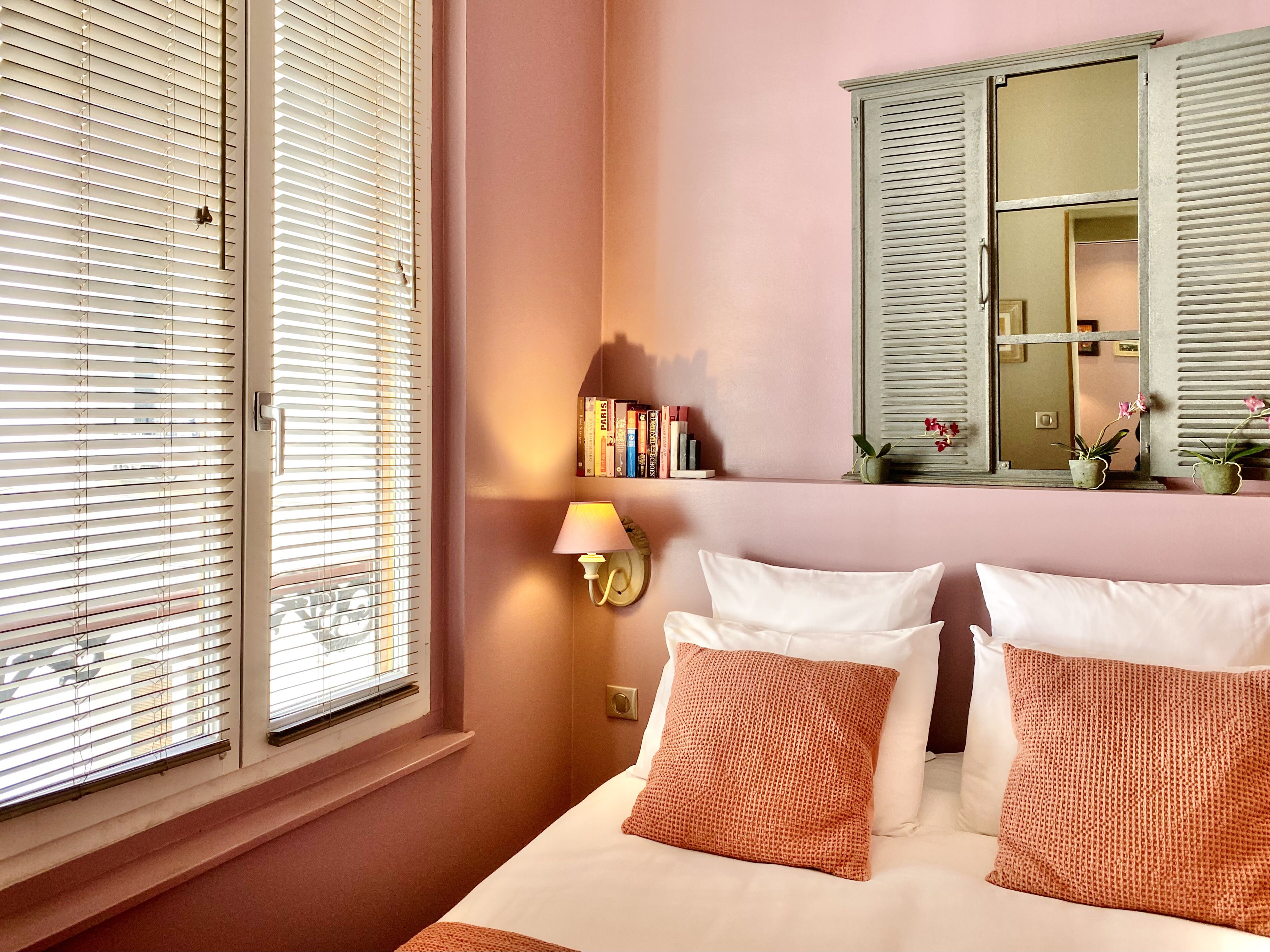 A bedroom with a bed, pillows, a nightstand, a lamp, a mirror, and a window with blinds.