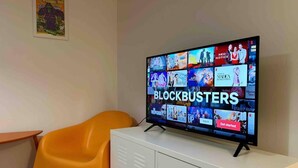 Een televisie toont een selectie films en tv-programma’s, waaronder ‘BLOCKBUSTERS’, met een witte kast en een oranje fauteuil in de kamer.