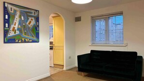 Een woonkamer met een donkergroene bank, een schilderij aan de muur, een raam met jaloezieën en een deur naar een keuken.