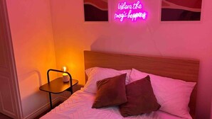 Een slaapkamer met een bed, kussens, een nachtkastje met een lamp en een neonbord aan de muur.