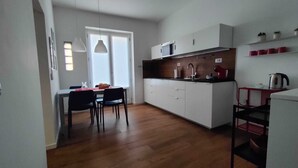 Een kleine keuken met witte kastjes, een magnetron, een spoelbak en een rode waterkoker. Er is een eethoek met een tafel en stoelen.