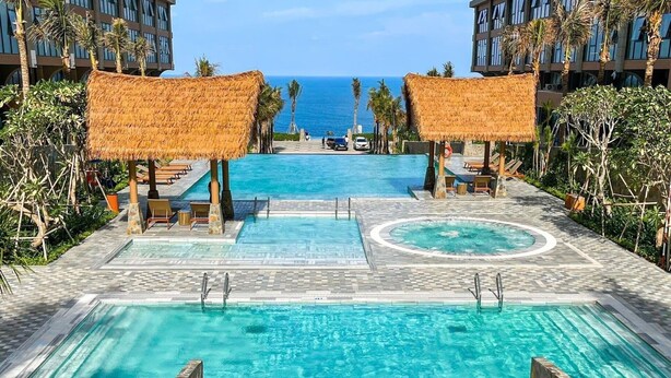 Ein Resort-Poolbereich mit mehreren Schwimmbecken, Liegestühlen und Reetdachschirmen.