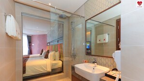 Ein modernes Hotelzimmer mit Duschkabine aus Glas, einem großen Bett mit farbigem Kopfteil, einem Badezimmer-Waschtisch aus Marmor und einem großen Spiegel.