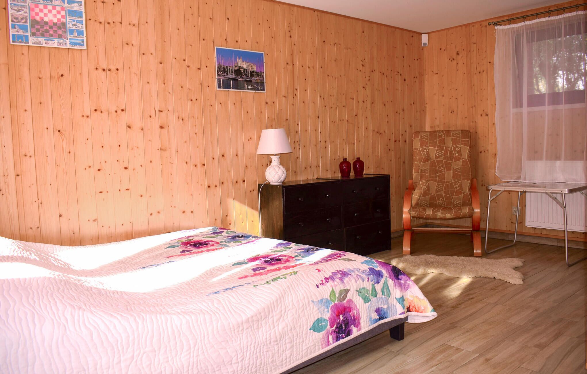 A bedroom with a wooden wall, a bed with a floral bedspread, a dresser, a chair, and a window with sheer curtains.