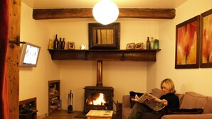 Un salon chaleureux avec une cheminée, des poutres en bois, un téléviseur et une personne qui lit un journal.