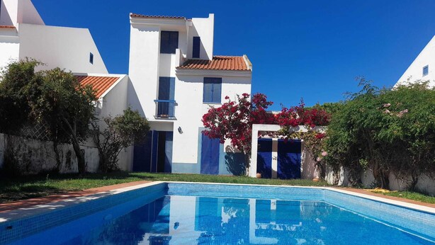 Una piscina con superficie de agua azul cristalina, rodeada de edificios blancos con contraventanas azules y flores rojas.