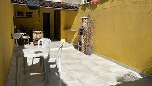 Eine patioartige Sitzgelegenheit mit gelben Wänden und Fliesenboden, einem weißen Plastiktisch und Stühlen, einem Korbstuhl und einer kleinen, aus Ziegeln gemauerten Struktur mit einer Pflanze.