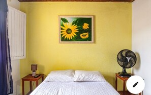 A bedroom with a bed, a nightstand with a lamp, a wall-mounted fan, and a painting of sunflowers.