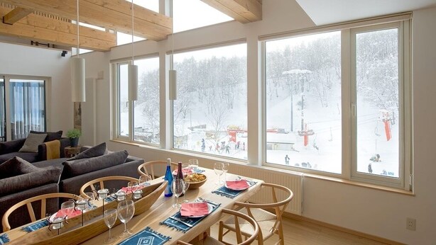 A dining area with a wooden table set for four, large windows with a snowy landscape view, and a cozy seating area with a sofa and chairs. - Chalet Murasaki (Kutchan)