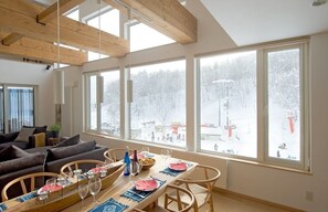 A dining area with a wooden table set for four, large windows with a snowy landscape view, and a cozy seating area with a sofa and chairs. - Chalet Murasaki (Kutchan)