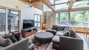 A modern living room with a large TV, comfortable seating, and large windows offering a view of the outdoors. - Chalet Murasaki (Kutchan)