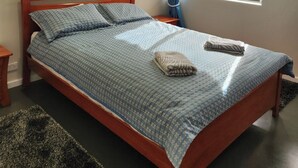 Siististi pedattu sänky, jossa on sininen kuviollinen päiväpeite ja tyynyjä, kattotuuletin, seinällä oleva metsäaiheinen taulu ja ikkuna, jossa on verhot.