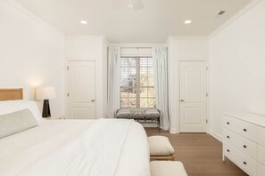 A bedroom with a large bed, a bedside table with a lamp, a window with curtains, and a bench.