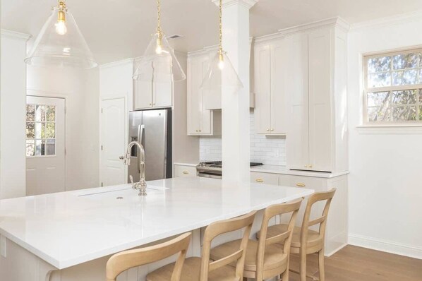A modern kitchen with white cabinetry, a central island with chairs, pendant lighting, and a stainless steel refrigerator.