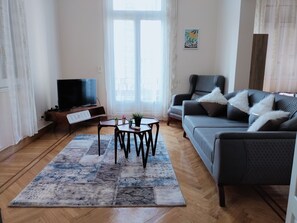 Uma sala de estar com sofá, mesa de centro, TV e janela com cortinas.