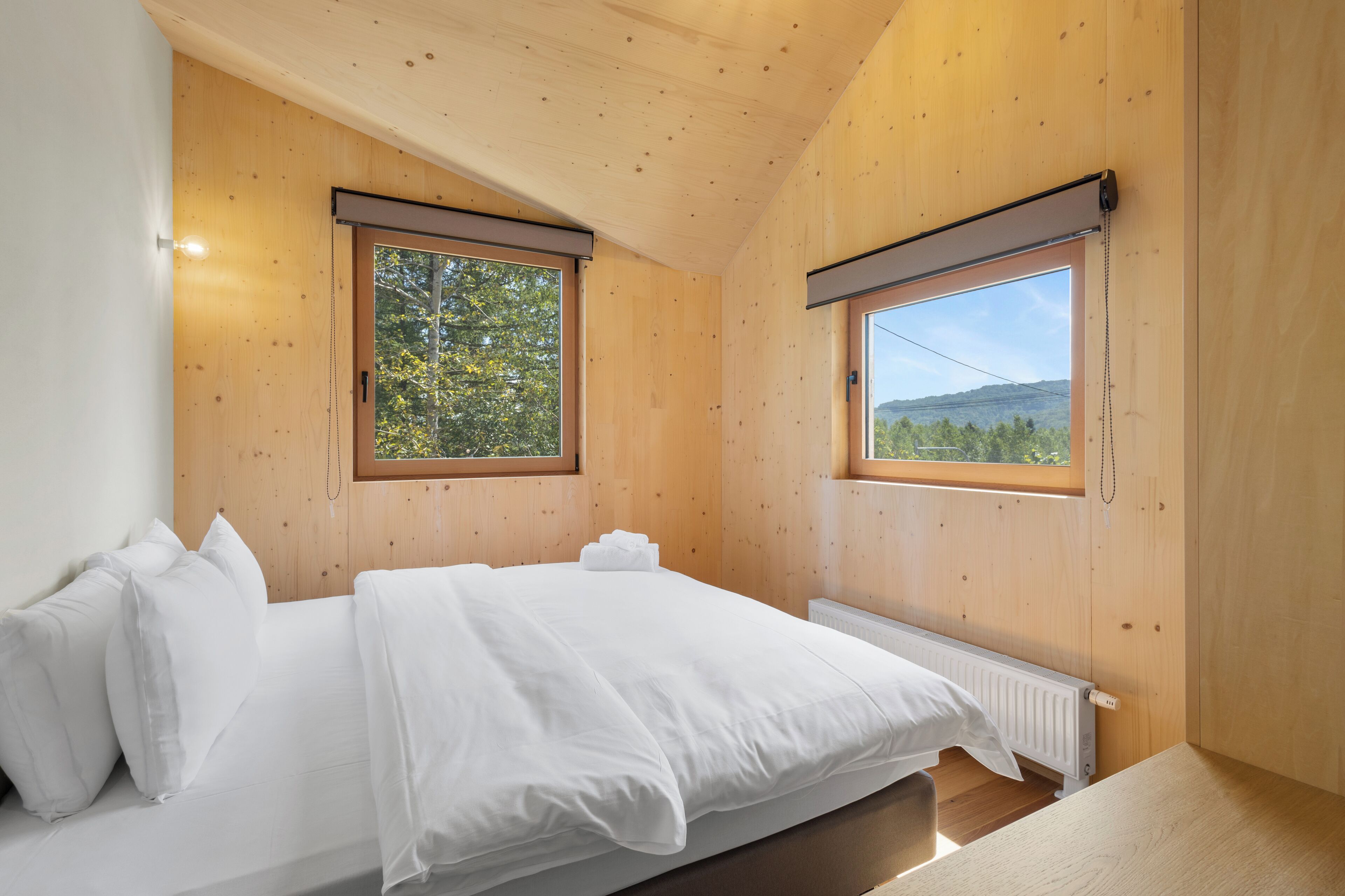 A bedroom with a wooden ceiling, a large bed with white bedding, two windows with blinds, and a view of trees and hills.