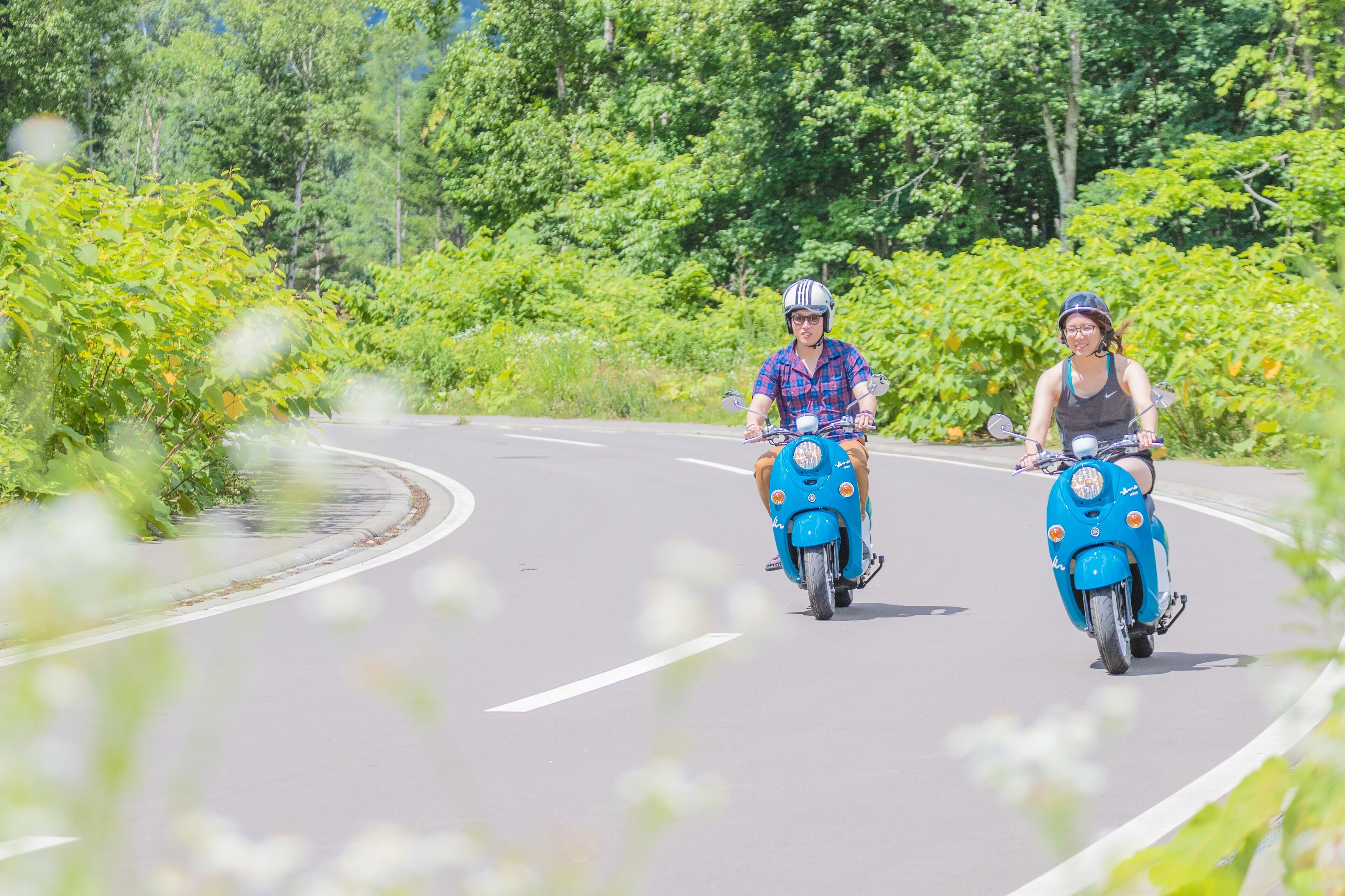 Two people riding blue scooters on a curved road surrounded by greenery.