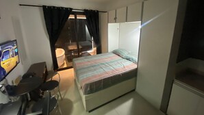 Quarto de hotel pequeno com uma cama, escrivaninha, cadeira, mesa pequena, televisão e janela com cortinas.