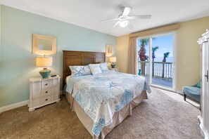 Ein Schlafzimmer mit Bett, Nachttischen, Deckenventilator, großem Fenster mit Blick auf Palmen und den Ozean sowie einem Balkon.