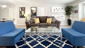 A modern living room with a grey sofa, two blue armchairs, a glass coffee table, and abstract wall art. - Muskegon Cozy Cutler Cottage. (Muskegon)