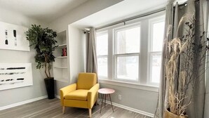 A modern living room with a yellow armchair, a small round table, a large window with curtains, a potted plant, and a bookshelf. - Muskegon Cozy Cutler Cottage. (Muskegon)