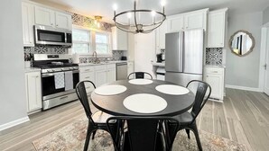 A modern kitchen with white cabinets, stainless steel appliances, and a round dining table with black chairs. - Muskegon Cozy Cutler Cottage. (Muskegon)
