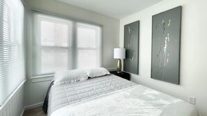 A bedroom with a bed, white bedding, a nightstand with a lamp, and two abstract paintings on the wall. - Muskegon Cozy Cutler Cottage. (Muskegon)