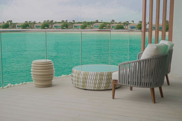 A balcony with a view of turquoise water, a round table, and a wicker chair.