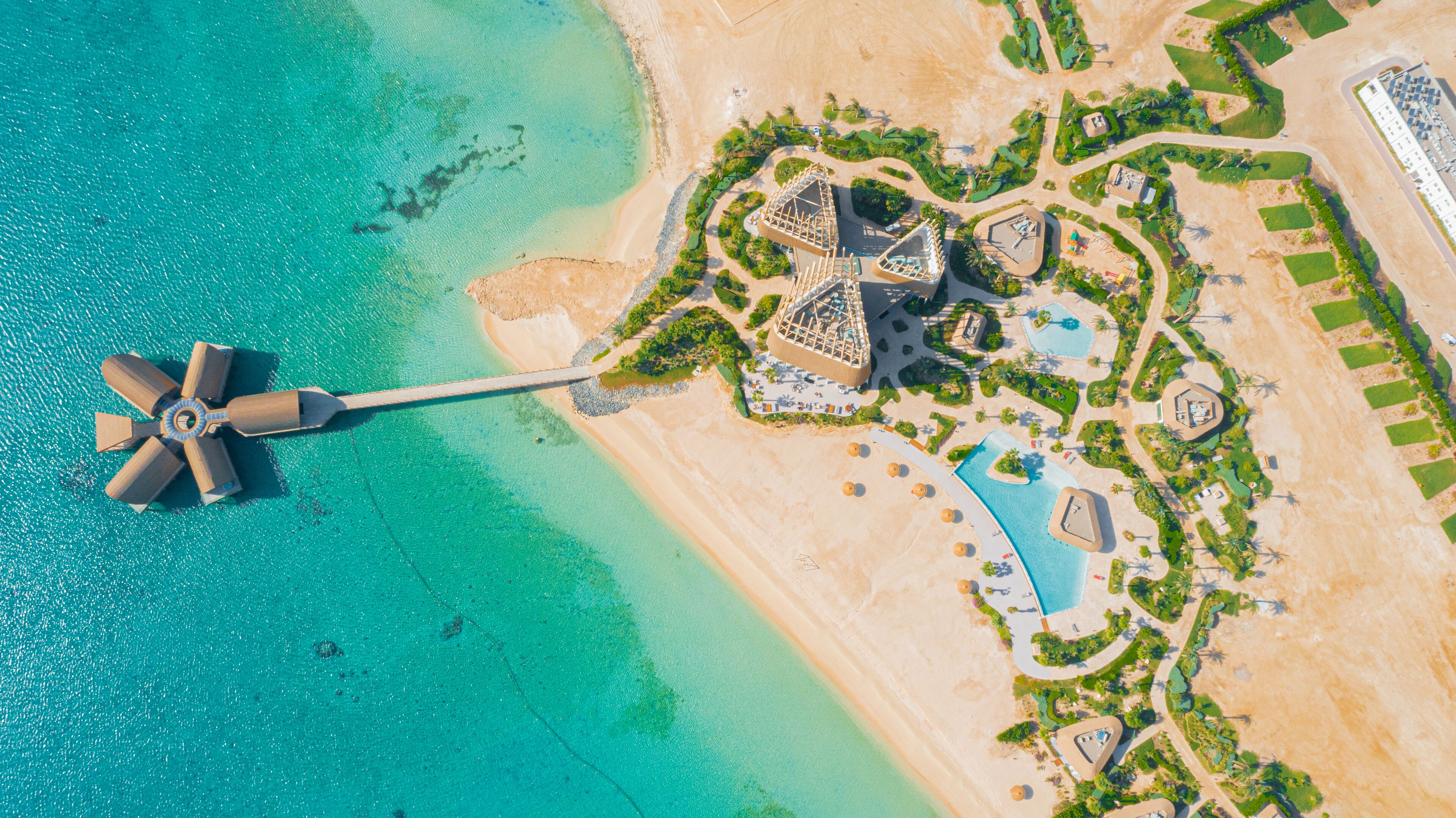 Vista aérea de um resort com praia, piscina e uma grande estrutura de ventoinha.