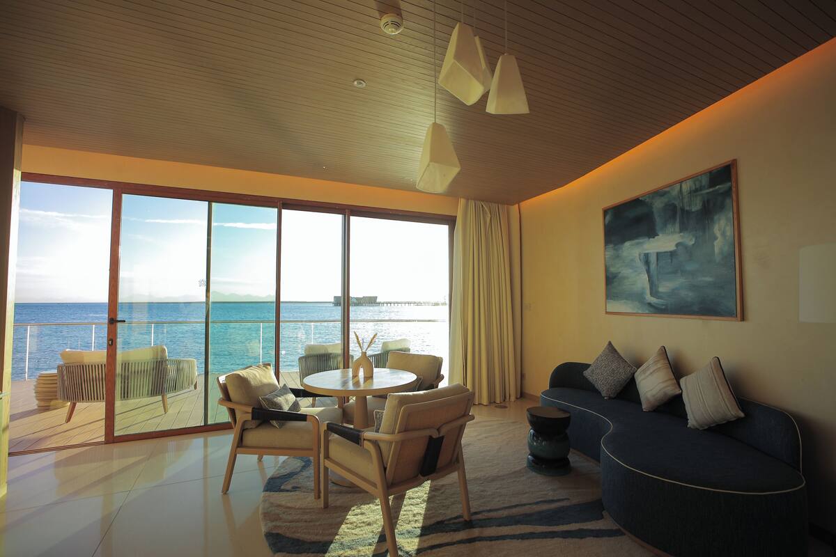 A spacious living room with a large sliding glass door leading to a balcony overlooking the sea. There is a round table with chairs, a sofa, and a painting on the wall.