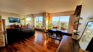 Un espacio habitable amplio con piso de madera, un sofá negro, una mesa de comedor con sillas y ventanas grandes que ofrecen vistas a la ciudad.