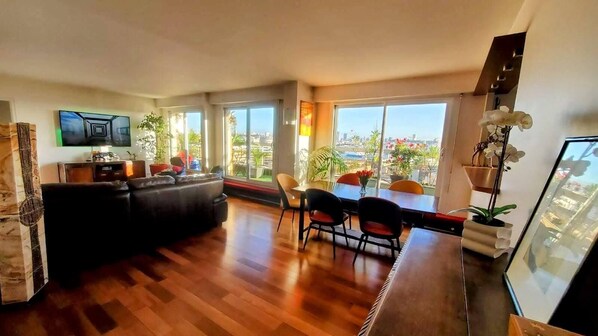 A spacious living area with a wooden floor, a black sofa, a dining table with chairs, and large windows offering a city view.