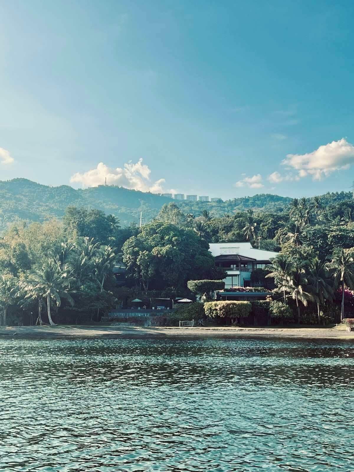 A waterfront property with a traditional building, lush greenery, and a view of distant hills.
