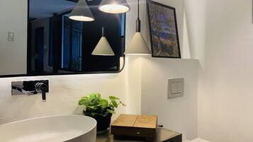Un baño moderno con un lavamanos grande blanco, un espejo, dos lámparas colgantes, una planta en maceta y un cuadro en la pared.