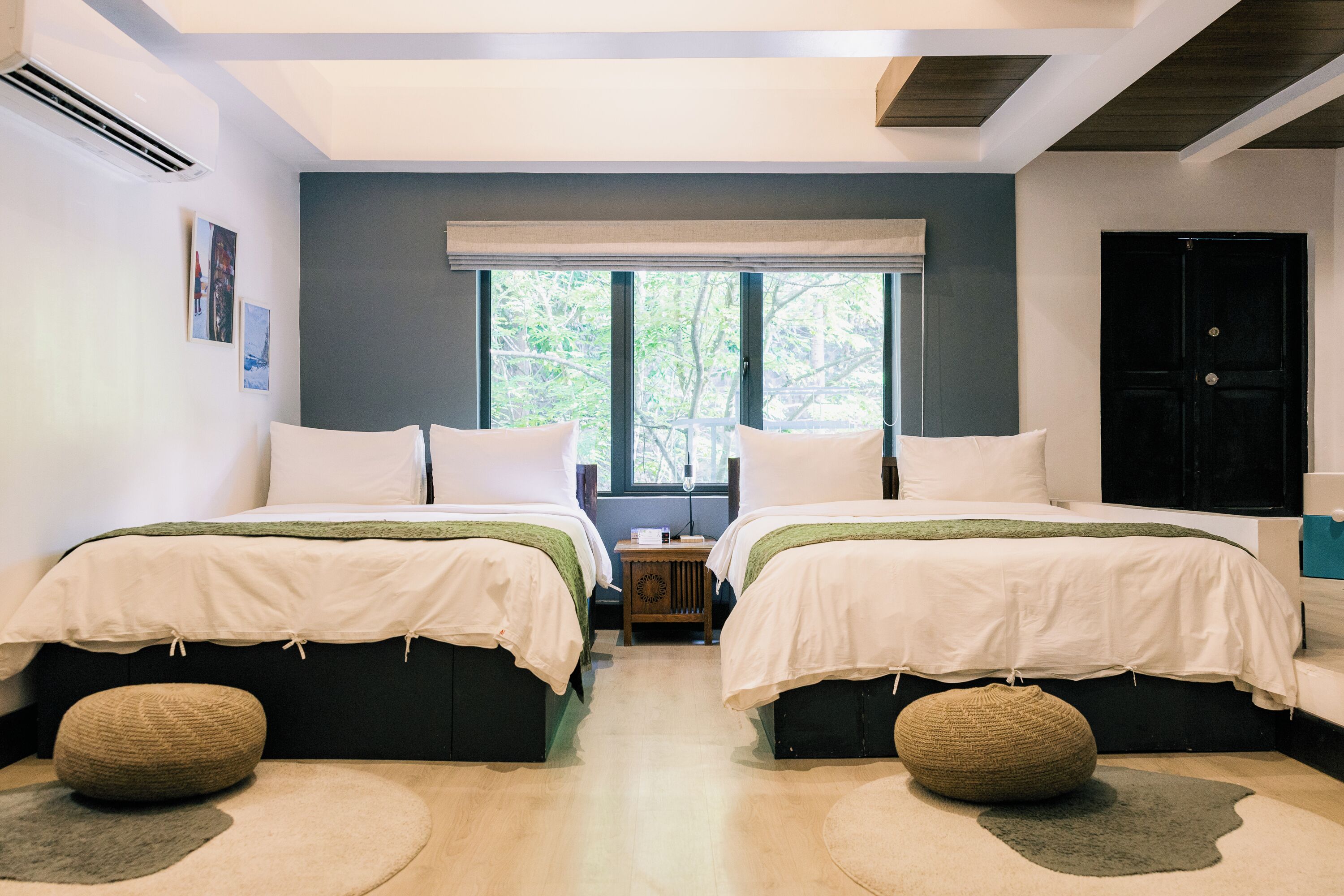 A modern bedroom with two beds, a large window, and a view of greenery outside.