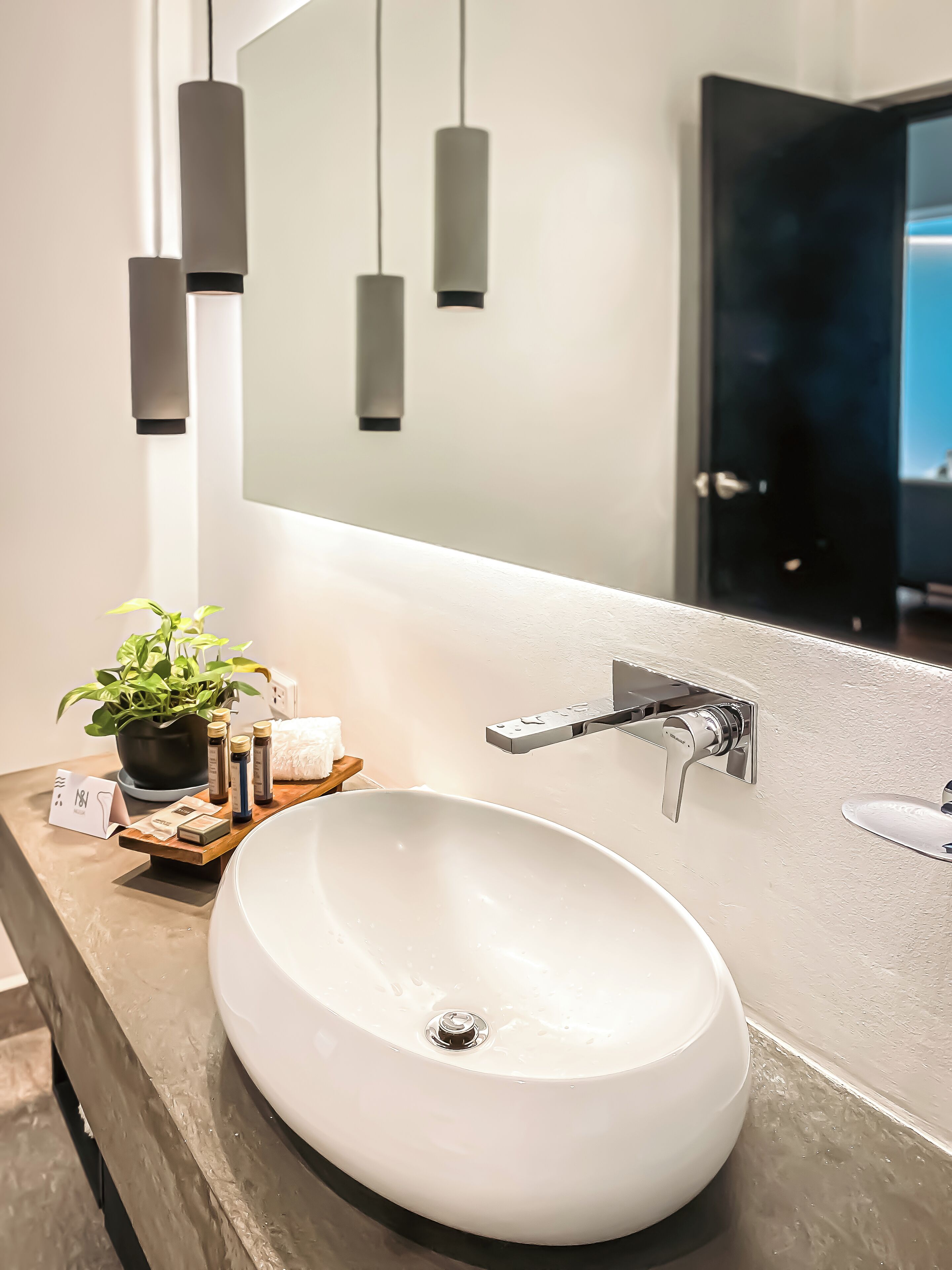 A modern bathroom with a white sink, a potted plant, and a mirror reflecting a dark room.