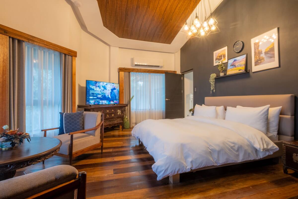 A modern hotel room with a large bed, wooden flooring, a television, a desk, and a chair.