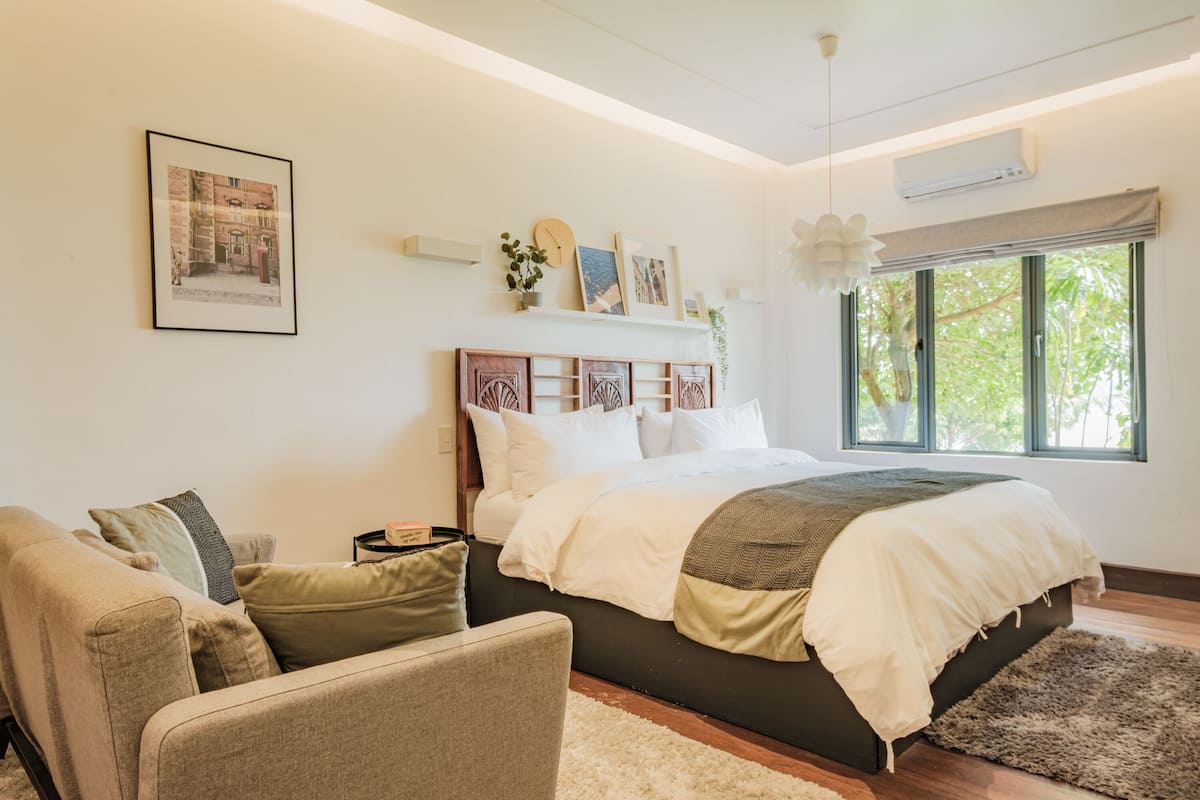 A bedroom with a bed, a sofa, a bedside table, and a window with a view of trees.