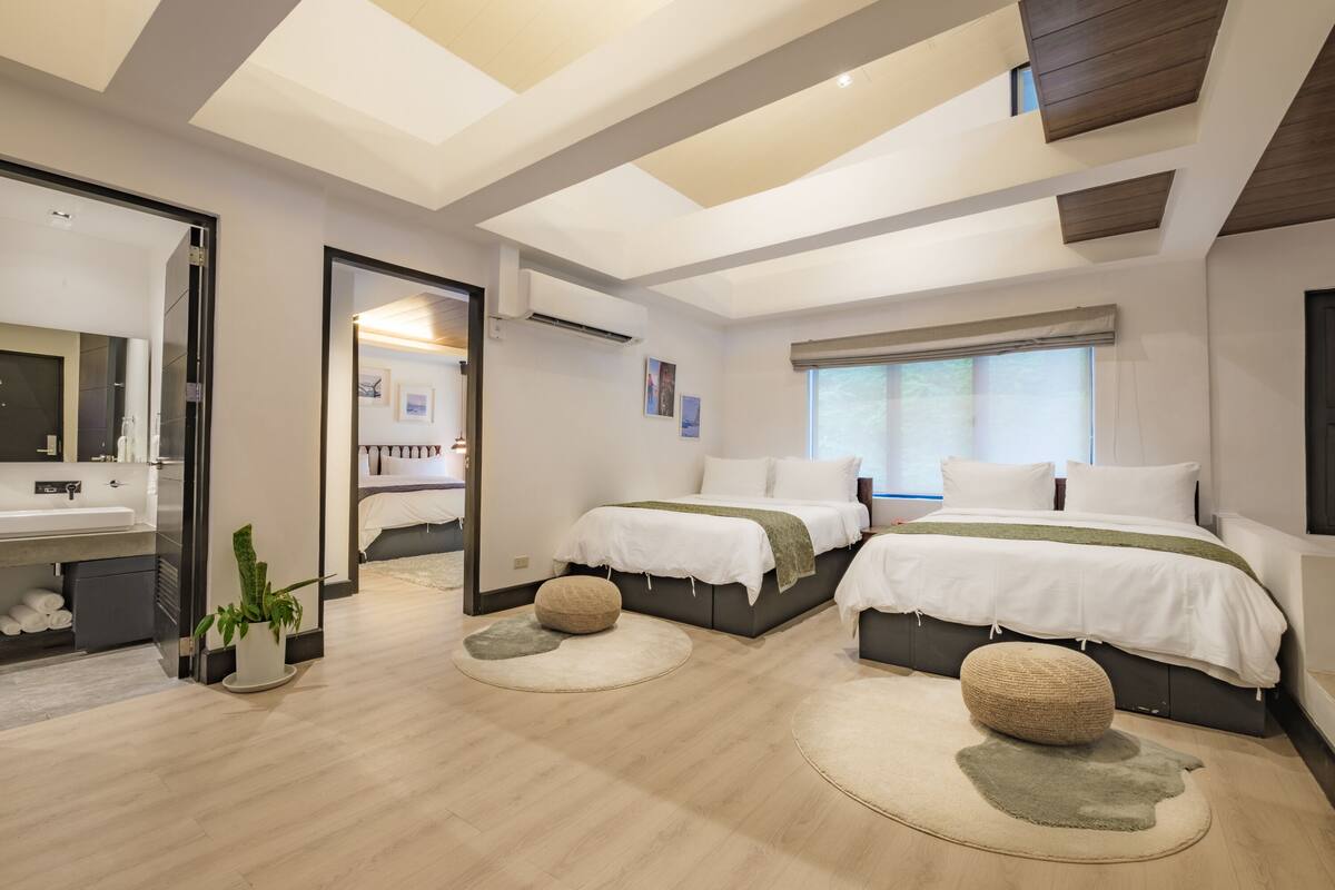 A modern bedroom with two beds, a large mirror, a bathroom with a sink, and a window providing natural light.
