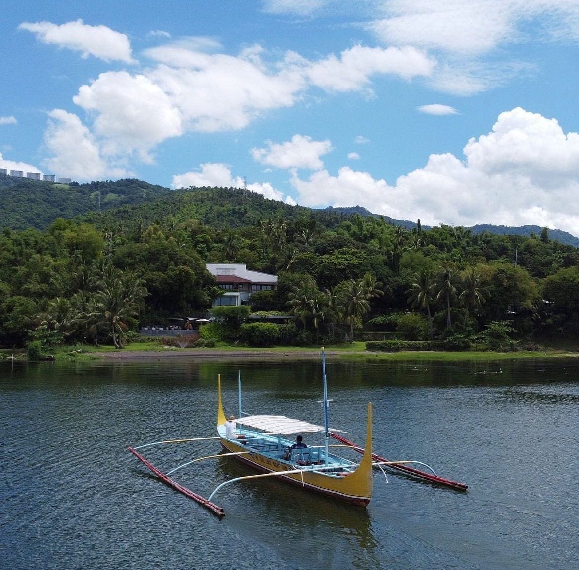 A traditional boat on a calm lake with a backdrop of lush greenery and a house on a hill.