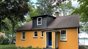 Une maison à deux étages, à la façade jaune et au toit foncé, avec une porte et des fenêtres bleues.